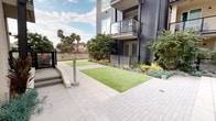 This outdoor courtyard serves as a central communal amenity within a contemporary multi-story residential building. The space is defined by its clean, modern aesthetic, combining wide light-toned paver walkways with expansive stretches of vibrant artificial turf that provide a low-maintenance yet visually inviting green environment. Thoughtfully curated landscaping beds line the perimeter, featuring a diverse mix of ornamental grasses, succulents, agaves, and native-style plantings that add texture and color throughout the year. Towering palm trees are visible beyond the fencing, lending a distinctly California character to the surroundings. The courtyard is framed on multiple sides by the building's contemporary façade, which incorporates a mix of dark corrugated metal paneling, smooth stucco, and large glass-railed balconies that extend from upper-floor residences. Outdoor seating arrangements, including rattan chairs and wooden accent tables, are positioned on the turf to create relaxed gathering spots that encourage social interaction. Accessible ramp features and wide concrete pathways ensure ease of movement throughout the space. The open-sky setting allows abundant natural light to flood the courtyard, creating a bright and welcoming atmosphere ideal for both relaxation and community engagement.