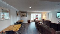 This single-floor space offers a well-appointed open layout that seamlessly blends relaxation and casual dining. The room features rich, dark hardwood flooring that grounds the space and contrasts beautifully with the neutral gray walls and textured ceiling. Recessed lighting is evenly distributed throughout, providing consistent, practical illumination complemented by a floor lamp near the seating area.

A large bay window fitted with white plantation shutters serves as a focal point, allowing natural light to filter into the room while maintaining privacy. Beneath and beside the window, a charming red popcorn machine adds a playful, entertainment-oriented accent, hinting at the room's leisure function.

The seating area is anchored by a row of brown reclining theater-style chairs arranged to face a focal viewing direction, making it ideal for a home cinema experience. Crown molding trims the upper walls, adding a refined architectural detail that elevates the overall finish of the space.

To the left, a casual dining nook features a natural wood table paired with metal bistro-style chairs, providing a functional counterpoint to the lounge area. Framed artwork adorns the walls, contributing to a warm, lived-in ambiance throughout.