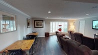 This single-floor space offers a well-appointed open layout that seamlessly blends relaxation and casual dining. The room features rich, dark hardwood flooring that grounds the space and contrasts beautifully with the neutral gray walls and textured ceiling. Recessed lighting is evenly distributed throughout, providing consistent, practical illumination complemented by a floor lamp near the seating area.

A large bay window fitted with white plantation shutters serves as a focal point, allowing natural light to filter into the room while maintaining privacy. Beneath and beside the window, a charming red popcorn machine adds a playful, entertainment-oriented accent, hinting at the room's leisure function.

The seating area is anchored by a row of brown reclining theater-style chairs arranged to face a focal viewing direction, making it ideal for a home cinema experience. Crown molding trims the upper walls, adding a refined architectural detail that elevates the overall finish of the space.

To the left, a casual dining nook features a natural wood table paired with metal bistro-style chairs, providing a functional counterpoint to the lounge area. Framed artwork adorns the walls, contributing to a warm, lived-in ambiance throughout.