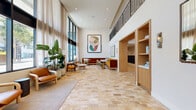 Located at 1430 Lincoln Blvd in Santa Monica, California, this thoughtfully designed lobby hallway makes a striking first impression with its dramatic double-height ceilings and expansive floor-to-ceiling windows. Natural light pours in through the large glazed entrance facade and flanking window walls, illuminating the warm, patchwork stone tile flooring that anchors the entire space with texture and character.

The entry zone features a set of floor-to-ceiling steel-framed glass doors that open to the exterior, framing views of the surrounding streetscape and lush tropical plantings arranged in oversized vessels nearby. Sheer floor-length drapery softens the abundant natural light along the window walls, lending an airy, refined atmosphere.

Moving deeper into the hallway, a generously sized lounge area is furnished with rich velvet banquette seating in deep rust and amber tones, paired with sculptural wooden chairs and rounded marble-topped tables. Bold, large-format abstract artwork adorns the white walls, adding a vibrant artistic dimension. An upper-level mezzanine with a railing overlooks the space below, reinforcing the impressive vertical scale. Built-in wooden millwork with open shelving adds warmth, while recessed lighting complements the natural illumination throughout. The overall ambiance is welcoming, contemporary, and distinctly California-inspired.