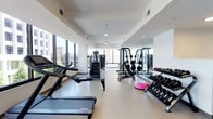 This fitness center offers a thoughtfully designed workout environment within a clean, contemporary setting. The space is characterized by crisp white walls, smooth light-toned flooring, and a bright, open atmosphere enhanced by large windows along multiple walls that provide abundant natural light and outward views of the surrounding urban landscape. Ceiling fans and recessed lighting complement the natural illumination throughout.

The cardio zone is well-stocked with treadmills, elliptical machines, stationary bikes, and a stair climber, all positioned to take advantage of the expansive window views. A wall-mounted flat-screen television is conveniently located nearby for entertainment during workouts. The strength training area features a multi-station cable machine, a squat rack loaded with weight plates, a dumbbell rack with a full range of free weights, an adjustable bench, and various accessories including balance trainers. Floor-to-ceiling mirrors line one wall, visually expanding the space and allowing users to monitor their form.

A separate corridor leads to additional amenity rooms, and the entrance area is accented with vibrant artwork and lush potted plants, adding a welcoming touch to the overall functional design.