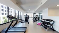 This fitness center offers a thoughtfully designed workout environment within a clean, contemporary setting. The space is characterized by crisp white walls, smooth light-toned flooring, and a bright, open atmosphere enhanced by large windows along multiple walls that provide abundant natural light and outward views of the surrounding urban landscape. Ceiling fans and recessed lighting complement the natural illumination throughout.

The cardio zone is well-stocked with treadmills, elliptical machines, stationary bikes, and a stair climber, all positioned to take advantage of the expansive window views. A wall-mounted flat-screen television is conveniently located nearby for entertainment during workouts. The strength training area features a multi-station cable machine, a squat rack loaded with weight plates, a dumbbell rack with a full range of free weights, an adjustable bench, and various accessories including balance trainers. Floor-to-ceiling mirrors line one wall, visually expanding the space and allowing users to monitor their form.

A separate corridor leads to additional amenity rooms, and the entrance area is accented with vibrant artwork and lush potted plants, adding a welcoming touch to the overall functional design.