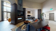 This thoughtfully designed single-floor amenity space blends contemporary style with a vibrant, social atmosphere. The layout flows seamlessly across several distinct zones, each offering its own character while contributing to a cohesive overall aesthetic.

The main lounge area anchors the space with soaring ceilings, a dramatic brick-accented fireplace framed by dark wood paneling, and a curved sectional sofa positioned around a sculptural round coffee table. An abstract area rug and curated wall art, including a striking figure silhouette, add artistic depth to the room.

Adjacent to the lounge, a dedicated game room features a pool table with a statement chandelier overhead, creating an inviting social hub. A separate area houses a shuffleboard table set beneath a gallery wall of framed artwork celebrating music and culture. A card or game table with plaid upholstered chairs and a mounted flat-screen TV rounds out the entertainment offerings.

The open corridor and entry areas are finished with large-format porcelain tile flooring and feature eye-catching geometric patterned accent walls in black and white, lending a bold visual identity to the transitional spaces. Rattan bistro seating, colorful wall-mounted display boxes, and ample natural light streaming through floor-to-ceiling windows create a welcoming, gallery-like atmosphere throughout.