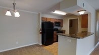 This single-floor space showcases an open-concept layout that connects the kitchen and living area, creating a cohesive and functional flow throughout. The kitchen is equipped with black appliances, including a full-size refrigerator, gas range, and built-in microwave, all complemented by warm medium-toned wood cabinetry above. Dark granite-style countertops provide a contrasting accent, and a peninsula with a breakfast bar extends into the living space, offering additional prep or dining space. The kitchen features light hardwood-style flooring, while the adjacent living area transitions to carpeting, subtly defining each zone. Crown molding runs along the upper walls, adding a clean architectural detail to the space. The textured ceiling and neutral beige walls create a simple, understated backdrop. A doorway visible at the rear of the kitchen suggests additional rooms beyond, indicating a practical layout with clear separation between living and private spaces. Overall, the space presents a straightforward, functional design with an easy-to-maintain interior that balances comfort and utility on a single level.