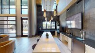 Located at 930 Jones Franklin Rd in Raleigh, NC, this single-floor amenity space delivers a sophisticated blend of modern design and inviting communal functionality. The centerpiece of the space is a dramatic double-height lounge area enclosed by floor-to-ceiling gridded windows, flooding the interior with natural light and offering expansive outdoor views. Oversized circular chandeliers and light wood flooring complement the warm, neutral tones of leather lounge seating arranged around coffee tables, creating an ideal setting for relaxed gatherings.

A fully equipped kitchen and bar area features a lengthy white quartz island with barstool seating, dark granite countertops, white cabinetry, stainless steel appliances, a draft beer tap, and striking oxidized metal wall panels. Pendant Edison-bulb fixtures hang from the soaring ceiling above, adding an industrial-chic character to the space.

A staircase with metal cable railings leads to an upper mezzanine level, with decorative woven leaf wall art adding an organic, artistic touch to the double-height wall. Adjacent areas include a game room outfitted with classic arcade machines and a foosball table, accented by reclaimed wood ceiling panels and built-in shelving. Glass-panel doors connect the various zones, maintaining visual openness throughout.