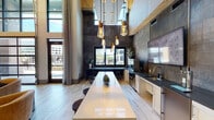 Located at 930 Jones Franklin Rd in Raleigh, NC, this single-floor amenity space delivers a sophisticated blend of modern design and inviting communal functionality. The centerpiece of the space is a dramatic double-height lounge area enclosed by floor-to-ceiling gridded windows, flooding the interior with natural light and offering expansive outdoor views. Oversized circular chandeliers and light wood flooring complement the warm, neutral tones of leather lounge seating arranged around coffee tables, creating an ideal setting for relaxed gatherings.

A fully equipped kitchen and bar area features a lengthy white quartz island with barstool seating, dark granite countertops, white cabinetry, stainless steel appliances, a draft beer tap, and striking oxidized metal wall panels. Pendant Edison-bulb fixtures hang from the soaring ceiling above, adding an industrial-chic character to the space.

A staircase with metal cable railings leads to an upper mezzanine level, with decorative woven leaf wall art adding an organic, artistic touch to the double-height wall. Adjacent areas include a game room outfitted with classic arcade machines and a foosball table, accented by reclaimed wood ceiling panels and built-in shelving. Glass-panel doors connect the various zones, maintaining visual openness throughout.
