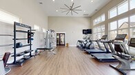 This well-appointed fitness center is designed to accommodate a comprehensive workout experience within a single open floor plan. The space is immediately distinguished by its dramatically high ceilings, which lend an open, airy atmosphere and are complemented by a large industrial-style ceiling fan that promotes airflow throughout the room. Recessed lighting is integrated throughout, ensuring consistent illumination across the entire facility.

Floor-to-ceiling windows line multiple walls, flooding the interior with natural light and offering views of the surrounding community, including a pool area visible from the cardio zone. The warm hardwood-style flooring runs throughout, providing both durability and a clean aesthetic.

The cardio section is well-equipped with multiple treadmills, elliptical trainers, and stationary bikes, all positioned to face the windows and a wall-mounted flat-screen television. On the opposite side of the space, a dedicated strength training area includes a multi-functional cable machine, a Body-Solid free weight rack, adjustable benches, kettlebells, medicine balls, and a dumbbell rack. A double-door entry provides easy access, and the neutral, warm-toned walls contribute to a clean and motivating environment.