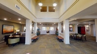 This single-story community clubhouse at Affinity at Lacey is designed to offer residents a comprehensive range of amenities within a cohesive, well-appointed environment. The grand entry lobby greets visitors with soaring ceilings, recessed lighting, warm neutral tones, patterned carpet tile flooring, flanking console tables, decorative potted plants, and framed artwork that set an inviting tone throughout.

The expansive central lounge features structural columns defining open seating zones furnished with bold orange accent chairs, leather armchairs, and coffee tables arranged around a fireplace. Large windows allow natural light to filter throughout, creating a bright, airy atmosphere. Adjacent areas provide additional casual seating with a wall-mounted television for gathering and relaxation.

The dedicated business center is equipped with multiple computer workstations, a printer, and a coffee station, all set against hardwood-style flooring and vibrant framed artwork. Dual stainless-steel water fountains are conveniently located nearby.

The private screening room is a standout feature, offering tiered rows of plush leather theater-style seating with cup holders, a large projection screen, wall sconce lighting, a striped carpet, and a compact refreshment bar with a retro-style mini refrigerator — providing a true cinematic experience within the community.