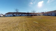 This single-floor unit is part of a well-maintained, low-rise residential complex set against an open, grassy landscape. The exterior presents a row of white-sided buildings with low-pitched roofs, set back from a generous open lawn that provides a sense of space and openness under wide, sunny skies.

Upon entering through the numbered front door, a bright entryway greets residents with large floor-to-ceiling windows that flood the space with natural light and frame views of the surrounding grounds. The interior features neutral, white-painted walls and warm wood baseboards throughout, creating a clean and cohesive aesthetic.

The main living area is generously proportioned, with patterned wall-to-wall carpeting, a ceiling fan with integrated light fixture, and multiple windows that allow ample natural light. The open layout connects seamlessly to adjacent spaces, including a closet and a kitchenette or kitchen area visible through a doorway, fitted with warm wood cabinetry.

Additional rooms mirror the same neutral palette and carpeted flooring, each equipped with windows that maintain the sense of light and connection to the outdoors. Floor vents, standard electrical outlets, and clean wall finishes reflect practical, functional design throughout. The overall ambiance is bright, quiet, and move-in ready, well-suited for comfortable single-level living.