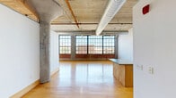 Located at 2034 Lucas Ave in St. Louis, MO, this loft-style unit on the fourth floor offers a compelling blend of industrial character and modern functionality. The open main living area is defined by soaring ceilings with exposed concrete and wood decking, visible steel support columns, and large-scale industrial windows that line an entire wall, flooding the space with abundant natural light and framing views of the surrounding St. Louis cityscape. Polished hardwood floors run throughout, complementing the raw architectural elements above. Exposed silver ductwork and track lighting add to the unit's distinctly industrial aesthetic, while crisp white walls provide a clean, versatile backdrop. A plywood accent wall introduces a warm, natural texture that softens the space. The kitchen is thoughtfully appointed with light maple cabinetry, solid countertops, stainless steel appliances including a French door refrigerator and electric range with overhead microwave, and durable hardwood flooring that carries through from the main living area. A dedicated utility room provides practical storage and laundry solutions. Steel structural columns are integrated throughout the open floor plan, serving as architectural focal points that honor the building's industrial heritage.