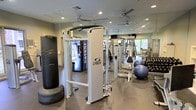 This fitness center is designed to support a comprehensive workout experience, offering a thoughtfully arranged layout across a single floor. The space is outfitted with professional-grade Cybex cable and strength training machines, providing users with versatile resistance training options. A heavy punching bag mounted near the entrance adds a functional combat fitness element, while a rack of dumbbells and stability balls accommodates free weight and core training needs. Full-length mirrors line the walls, visually expanding the space and allowing users to monitor their form throughout their workout. The dark rubberized flooring provides both safety and durability underfoot. Ceiling fans are distributed throughout, promoting air circulation and comfort during exercise. Recessed lighting ensures the space is evenly and adequately illuminated. A Dasani water vending machine is conveniently positioned toward the back, offering hydration access during sessions. Natural light filters in through a window visible at the rear of the space, contributing to an open and energizing atmosphere. The overall design balances functionality and comfort, making it suitable for a wide range of fitness routines.