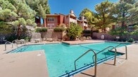Located in El Paso, Texas, this outdoor community pool area offers a well-maintained and inviting amenity space set against a backdrop of mature pine trees and classic brick architecture. The centerpiece is a generously sized swimming pool with clearly marked depth gradations, entry handrails, and a broad surrounding deck that provides ample room for lounging and socializing. Multiple seating arrangements featuring tables and chairs are distributed across the pool deck, offering comfortable spots for residents to relax under the abundant West Texas sunshine. A dedicated pool house constructed of traditional red brick anchors one end of the space, providing utility and visual structure to the area. Retaining walls of natural stone define the tiered landscaping, adding texture and a sense of depth to the surroundings. Lush, mature trees frame the entire area, offering both shade and privacy while contributing to the serene, resort-like atmosphere. The multi-story residential buildings surrounding the space feature balconies and chimneys, adding architectural character to the overall setting. The combination of open sky, verdant greenery, and warm brick tones creates a cohesive and welcoming outdoor environment ideal for community gathering and recreation.