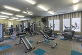This fitness center in Gallatin, Tennessee offers a comprehensive workout environment designed to accommodate a wide range of exercise needs. The single-level space features neutral gray walls paired with a patterned carpet floor accented by teal exercise mat sections, creating a functional yet visually cohesive aesthetic.

The room is well-lit through a combination of overhead fluorescent panel lighting and ceiling fans that promote air circulation throughout. Multiple windows fitted with linen-toned curtains and white blinds line the walls, allowing natural light to fill the space and contributing to an open, energizing atmosphere.

A full-length mirrored wall extends along one side of the room, visually expanding the space while supporting proper form during workouts. Equipment includes elliptical trainers, stationary bikes, treadmills, and a cable strength machine, all from recognized fitness brands. A dedicated free weight area features a multi-tiered dumbbell rack alongside stability and medicine balls, catering to strength and functional training needs.

A wall-mounted flat-screen television provides entertainment during workouts. The overall layout is thoughtfully organized, offering clearly defined zones for cardio and strength training while maintaining ample open floor space for stretching and floor-based exercises.
