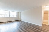 Located at 1257 Lakeshore Rd E in Mississauga, Ontario, this one-bedroom unit offers a thoughtfully organized layout across a single floor. The open living room is a standout feature, bathed in natural light through a wide, multi-panel window that frames views of mature trees and the surrounding neighbourhood. Rich hardwood-style flooring runs throughout the main living areas, complementing the neutral, warm-toned walls and clean white baseboards and crown moulding. Baseboard heating units run beneath the windows, providing efficient warmth throughout the space. The kitchen is well-equipped and galley-style in form, featuring warm wood cabinetry, granite countertops, a subway tile backsplash, stainless steel appliances including a refrigerator and range, and light tile flooring. A pass-through opening connects the kitchen to the living and dining area, enhancing the sense of openness. The generously sized bedroom accommodates comfortable furnishing arrangements and receives natural light through its own dedicated window with outdoor views. A closet with bi-fold doors provides practical storage. Crown moulding throughout adds a refined architectural touch, contributing to the clean, well-maintained character of the overall space.
