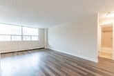 Located at 1257 Lakeshore Rd E in Mississauga, Ontario, this one-bedroom unit offers a thoughtfully organized layout across a single floor. The open living room is a standout feature, bathed in natural light through a wide, multi-panel window that frames views of mature trees and the surrounding neighbourhood. Rich hardwood-style flooring runs throughout the main living areas, complementing the neutral, warm-toned walls and clean white baseboards and crown moulding. Baseboard heating units run beneath the windows, providing efficient warmth throughout the space. The kitchen is well-equipped and galley-style in form, featuring warm wood cabinetry, granite countertops, a subway tile backsplash, stainless steel appliances including a refrigerator and range, and light tile flooring. A pass-through opening connects the kitchen to the living and dining area, enhancing the sense of openness. The generously sized bedroom accommodates comfortable furnishing arrangements and receives natural light through its own dedicated window with outdoor views. A closet with bi-fold doors provides practical storage. Crown moulding throughout adds a refined architectural touch, contributing to the clean, well-maintained character of the overall space.