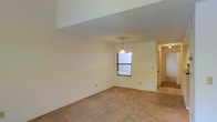 This two-floor residence offers a straightforward and functional layout designed for comfortable everyday living. The main living area features warm, neutral-toned carpeted flooring that extends throughout, providing a soft and cohesive feel underfoot. Smooth, light-colored walls create a bright and airy atmosphere, complemented by natural light filtering in through a window. A pendant light fixture hangs centrally, adding a subtle decorative touch to the space. The open layout allows for flexible furniture arrangement, making it easy to customize the area to suit various needs. A hallway leads to additional rooms and spaces, hinting at a well-organized floor plan that connects the various areas of the home seamlessly. The transition between the carpeted living space and what appears to be a tiled area near the hallway suggests a practical division between different functional zones within the home. With two floors, the property offers ample room for living, working, and storage, providing versatility for a range of lifestyles. The overall ambiance is clean, neutral, and move-in ready, with a blank-canvas aesthetic that invites personalization.