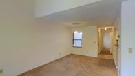 This two-floor residence offers a straightforward and functional layout designed for comfortable everyday living. The main living area features warm, neutral-toned carpeted flooring that extends throughout, providing a soft and cohesive feel underfoot. Smooth, light-colored walls create a bright and airy atmosphere, complemented by natural light filtering in through a window. A pendant light fixture hangs centrally, adding a subtle decorative touch to the space. The open layout allows for flexible furniture arrangement, making it easy to customize the area to suit various needs. A hallway leads to additional rooms and spaces, hinting at a well-organized floor plan that connects the various areas of the home seamlessly. The transition between the carpeted living space and what appears to be a tiled area near the hallway suggests a practical division between different functional zones within the home. With two floors, the property offers ample room for living, working, and storage, providing versatility for a range of lifestyles. The overall ambiance is clean, neutral, and move-in ready, with a blank-canvas aesthetic that invites personalization.