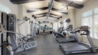 This fitness center is a spacious, single-floor facility designed to support a comprehensive workout experience. The space is defined by its striking vaulted ceiling adorned with bold dark exposed beams, which lend an architectural character uncommon in typical gym environments. Ceiling fans are mounted overhead, complementing the modern linear lighting suspended from the ceiling structure, while exposed HVAC ductwork adds an industrial accent.

Natural light flows generously through multiple large windows dressed with slatted blinds, offering views of lush greenery outside and creating a bright, welcoming atmosphere. The flooring throughout is a durable dark rubberized material, ideal for heavy equipment use.

The facility is outfitted with a robust selection of Cybex-brand equipment, including multiple treadmills, elliptical cross-trainers, and stair climbers arranged along one side of the room. On the opposing side, strength training is well-supported with a cable machine, a multi-function weight station, and an adjustable bench. A dedicated free weights area features a full rack of dumbbells and a barbell with weight plates. Medicine balls and resistance training accessories round out the strength zone.

Full-length mirrors line one wall, visually expanding the space and allowing users to monitor their form. Multiple wall-mounted flat-screen televisions are positioned throughout for entertainment during workouts.