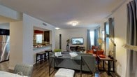This single-floor residence showcases a thoughtfully designed open-concept layout that seamlessly connects the kitchen and living areas. The living room is a welcoming, well-proportioned space anchored by a wall-mounted flat-screen television and furnished with a contemporary gray sectional sofa complemented by vibrant orange accent chairs. A sleek glass coffee table sits at the center, adding a modern touch. Natural light filters in through large windows dressed with floor-length gray curtains, enhancing the bright and airy ambiance of the room. A decorative flush-mount light fixture provides additional illumination overhead.

The kitchen is accessible via a pass-through opening, featuring a raised breakfast bar with dark stools ideal for casual dining. The kitchen presents dark cabinetry, stone-look countertops, and a tile backsplash, creating a stylish yet functional culinary space. A window above the sink allows natural light to brighten the area. The rich hardwood-style flooring flows consistently throughout, tying the spaces together cohesively and contributing to the warm, inviting character of the home.