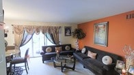 This single-floor space centers around a thoughtfully styled living room that makes a strong visual impression. The room features a bold terracotta-orange accent wall that serves as a warm focal point, complemented by neutral beige walls and light-colored carpeted flooring. Two dark charcoal upholstered sofas are arranged around a sleek, oval glass-top coffee table, creating a comfortable and inviting seating area. Decorative throw pillows in earthy tones and patterned fabrics add texture and visual interest throughout the space. A large sliding glass door dressed with ornate, layered drapery in rich gold and dark tones allows natural light to flow into the room while offering a connection to the outdoors. The walls are adorned with framed artwork, including a striking black-and-white portrait and a colorful floral landscape painting, adding a personal and artistic character to the space. A decorative tree with warm twinkling lights stands in the corner, contributing to the cozy ambiance. The smooth, white ceiling completes the room with a clean, open feel overhead. Overall, this space presents a warm, eclectic aesthetic with a mix of bold color choices and carefully selected decorative elements.
