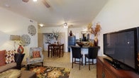 This single-floor residence offers an open-concept layout that effortlessly connects the living, dining, and kitchen areas into one cohesive and functional space. The living area is anchored by a ceiling fan and features warm neutral tones, complemented by a patterned area rug and comfortable seating. Large decorative accents and lush floral arrangements add character throughout the space.

The kitchen is equipped with white cabinetry, a stainless steel refrigerator, and dark-toned countertops. A breakfast bar with bar-height seating provides a convenient spot for casual dining, while the adjacent dining area accommodates a full dining set, making the space ideal for both everyday meals and entertaining.

Light tile flooring runs throughout the main living areas, creating a cohesive and easy-to-maintain surface. The ceiling fan centrally positioned above the dining and living zones ensures comfortable airflow throughout. The open sightlines between rooms create a sense of spaciousness and allow natural light to flow freely across the floor plan. The warm color palette, from the earthy tones of the décor to the neutral walls, gives the space a welcoming and lived-in ambiance that feels both stylish and practical.