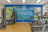 This fitness center is organized across two connected rooms, offering a comprehensive workout environment on a single level. The main exercise area features light hardwood-style flooring, vaulted ceilings, and large multi-pane windows that flood the space with natural light while providing views of the surrounding greenery. Ceiling fans assist with air circulation throughout. Cardio equipment, including treadmills and elliptical machines, lines the perimeter, accompanied by a dumbbell rack and kettlebell station. A wall-mounted flat-screen TV provides entertainment during workouts, and glass-paned double doors open to an outdoor area, enhancing the airy feel.

The secondary room connects seamlessly and is distinguished by a bold blue accent wall adorned with a motivational fitness-themed word mural. This space houses strength and resistance training machines, a stationary bike, and a weight bench. A large wall mirror expands the perceived depth of the room. Together, the two rooms provide a well-rounded facility suited for both cardiovascular training and strength conditioning, maintaining an energetic yet functional ambiance throughout.