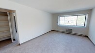 This single-floor unit presents a practical and functional layout across several rooms. The living areas are finished with neutral-toned wall-to-wall carpeting, white walls, and standard flat ceilings, creating a clean and understated aesthetic throughout. Multiple rooms benefit from natural light provided by windows fitted with vertical blinds, and each carpeted room is equipped with baseboard heating along with a wall-mounted air conditioning unit, ensuring year-round climate control.

The kitchen features wood-trimmed cabinetry in white and brown tones, offering ample upper and lower storage space. A double stainless steel sink, white coil electric range, and a full-size white refrigerator comprise the primary appliances. The kitchen is finished with hardwood-style flooring, distinguishing it from the carpeted rooms.

The bathroom is compact and efficiently arranged, featuring a full tub and shower combination with white square tile surround, a toilet, and a single vanity with a built-in sink and mirror. The bathroom floor is finished with hardwood-style planks, complementing the clean, simple design. Overall, the unit maintains a straightforward, functional character well-suited for everyday living.