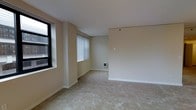 This single-floor space offers a straightforward, functional layout centered around an open living area. The room is finished with wall-to-wall carpeting in a neutral beige tone, providing a soft underfoot feel that complements the warm, cream-colored walls throughout. The smooth white ceiling adds a sense of openness to the space.

Natural light enters through multiple windows dressed with vertical blinds, allowing for adjustable light control and privacy. A smaller, darker-framed window provides an additional source of outside light, lending the space a connection to the outdoors. The windows are positioned to brighten the room while maintaining a sense of enclosure and comfort.

A wall-mounted heating unit is conveniently located along the baseboard, ensuring efficient climate control within the living area. The walls are largely unadorned, offering a blank canvas for personalization and furnishing. A subtle structural column adds a minor architectural element to the room's layout.

Overall, the space presents a clean, neutral, and move-in ready environment. Its open floor plan and understated design make it highly adaptable, suitable for a variety of furniture arrangements and interior styles. The combination of soft carpeting, natural light, and simple finishes creates a calm and practical living environment.