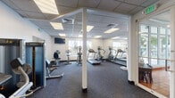 Located at 3875 Lombardy St in Hollywood, Florida, this single-story fitness and recreation facility offers a comprehensive layout designed to support a variety of exercise routines and athletic activities. The space is divided into two primary areas: a cardio and strength training gym and a full racquetball court.

The fitness room is bright and functional, featuring a drop ceiling with recessed panel lighting that ensures even illumination throughout. Large windows along one wall provide natural light and outdoor views of a pool area and tropical palm trees, creating an energizing workout environment. Full-length mirrors line one wall, visually expanding the space. Equipment includes treadmills, elliptical trainers, stationary bikes, and strength machines. A wall-mounted television adds convenience for users during cardio sessions. The carpeted flooring throughout the gym area provides comfort and stability underfoot.

Adjacent to the gym is a regulation racquetball court, distinguished by its polished hardwood flooring with clearly marked red boundary lines. The court features soaring ceilings that accommodate full gameplay, smooth white walls on all sides, and overhead lighting. A doorway connects the court directly to the fitness room, allowing seamless access between the two spaces. Together, these areas form a well-rounded recreational facility suited for diverse fitness needs.