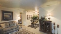 This single-floor residence offers a welcoming interior defined by a seamlessly connected living and dining area. The living space features plush, neutral-toned carpeting that extends throughout, contributing to a cozy and cohesive atmosphere. A textured ceiling with a ceiling fan provides both character and functional airflow overhead. The living area is anchored by a comfortable upholstered sofa, complemented by a classic side table with a lamp that casts a warm, ambient glow. Adjacent to the living space, the dining area is furnished with elegant wooden chairs and a round table set atop a richly patterned area rug, adding a refined touch to the interior. Decorative wall art and lush potted plants bring natural elements and visual interest to the walls, enhancing the overall warmth of the space. A dark wood display cabinet with glass-paneled doors stands prominently, offering both storage and decorative appeal. Soft, recessed lighting combined with warm lamp light creates an inviting ambiance throughout. A hallway visible from the main area suggests additional rooms beyond, rounding out this well-appointed, single-level home.