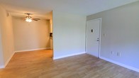 This single-floor residential space offers a straightforward and functional layout designed for comfortable everyday living. The main living area greets you at the entry door, featuring light hardwood-style flooring that extends throughout the space, lending warmth and continuity to the interior. The walls are finished in a neutral, soft gray-white tone, creating a clean and airy atmosphere that pairs well with a variety of furnishing styles. The entry door is framed with crisp white trim and includes a standard security lock, providing a welcoming and secure point of entry. A ceiling fan positioned in the adjacent area adds both practical ventilation and a subtle decorative touch to the space. An open passage connects the living area to the kitchen, which is visible from the main room. The kitchen features white cabinetry and countertops, maintaining the light and neutral aesthetic found throughout the unit. White baseboards run along the perimeter of the rooms, adding a clean, finished look to the overall interior. The single-floor design ensures ease of navigation throughout the space, making it an accessible and efficiently organized home. The combination of neutral tones, light flooring, and open sightlines gives the space a bright, uncluttered ambiance well-suited to a range of lifestyles.