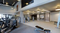 This fitness center offers a thoughtfully designed workout space with a contemporary aesthetic. The room features notably high ceilings with an elevated mezzanine-level window panel above, adding architectural depth and visual interest. Recessed and pendant lighting fixtures are arranged throughout the ceiling, providing bright, even illumination across the entire floor. The flooring consists of durable, light-colored carpet suitable for a fitness environment. Strength training equipment, including cable and weight machines, is arranged systematically across the floor, while cardio equipment such as an elliptical trainer is positioned toward the front. A dedicated adjustable workout bench adds versatility for free-weight exercises. Full-length mirrors line one wall, enhancing the sense of openness and allowing users to monitor their form. Decorative wall art and an exposed brick accent wall contribute warmth and character to the otherwise modern, neutral-toned interior. Structural columns with a textured finish add an industrial touch, complementing the clean white walls and gray tones throughout. The overall ambiance is polished and professional, creating a motivating environment for exercise.