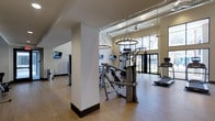 This two-floor fitness facility combines functionality with sophisticated design to create an inviting and well-equipped workout environment. The main exercise area features hardwood-style flooring that adds warmth and a polished aesthetic to the space, while structural columns provide architectural definition throughout the floor plan.

The facility is outfitted with a comprehensive selection of professional-grade strength and cardio equipment, including multi-use cable machines, treadmills, and a punching bag, catering to a wide range of fitness needs. Multiple flat-screen televisions are mounted throughout the space, allowing members to stay entertained during their workouts.

One of the standout design elements is the elegant circular chandelier-style lighting fixtures that hang from the high ceilings, adding an unexpected touch of refinement to the athletic setting. Exposed ductwork along the upper portion of the ceiling contributes an industrial character that complements the modern aesthetic.

Large floor-to-ceiling windows along the far wall flood the space with natural light and offer views of the outdoors, creating an open and energizing atmosphere. Recessed lighting further enhances the brightness of the interior. The overall ambiance of the facility is clean, contemporary, and thoughtfully designed to motivate and support an active lifestyle across both levels.