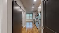 Located in Rogers, Arkansas, this single-floor unit offers a clean, contemporary design with warm hardwood-style flooring that flows throughout the entire space. The open-concept layout connects the kitchen and living area seamlessly, making the unit feel bright and airy.

The kitchen is well-appointed with gray shaker-style cabinetry, granite-look countertops, open wooden shelving, and a white subway tile backsplash. A full suite of stainless steel appliances — including a refrigerator, electric range, over-the-range microwave, and dishwasher — completes the space with a modern, functional finish.

The living area is generously sized, featuring neutral gray walls, recessed lighting, a ceiling fan, and multiple windows fitted with blinds that allow for natural light and privacy. A sliding barn door adds a stylish architectural detail, lending a contemporary-industrial character to the interior.

The bathroom is thoughtfully designed with warm wood-tone flooring continuing from the main living space, complemented by clean white tile in the walk-in shower area. The layout is practical and uncluttered. Recessed lighting throughout the unit ensures consistent, modern illumination in every area.