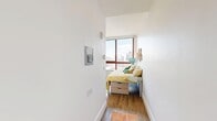 Located at 229 Duffield St in the heart of Downtown Brooklyn, this efficiently designed single-floor unit makes the most of every square inch. The layout includes a bedroom, bathroom, closet, and hallway, all thoughtfully arranged for functional living.

The bedroom is the focal point of the unit, anchored by a large floor-to-ceiling window framed in warm wood tones that floods the space with natural light and offers sweeping views of the Brooklyn skyline. Hardwood flooring runs throughout, adding warmth and continuity. A built-in workstation with shelving and storage drawers provides a dedicated workspace, while a compact refrigerator and microwave unit integrates kitchen convenience directly into the room. The bed features under-frame storage drawers, maximizing utility in the compact footprint.

The closet area is accessed via clean white double doors, offering organized storage space. The bathroom is finished with neutral tile walls and flooring, and includes an open-shelf vanity with a wall-length mirror, a toilet, and a step-in shower, all well-lit with overhead lighting. The hallway connects the spaces efficiently, maintaining an open and airy feel throughout. This unit is ideally suited for those seeking a smart, modern living space in a prime Brooklyn location.