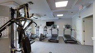 This fitness center offers a clean, well-organized workout environment designed to support a variety of exercise routines. The space is finished with neutral gray walls, crown molding trim, and a carpeted floor that lends a polished, comfortable feel. Bright recessed panel lighting in a coffered ceiling arrangement ensures the room is evenly and generously illuminated throughout.

The cardio section is prominently featured, housing multiple treadmills and elliptical trainers arranged in a row, each equipped with individual display consoles. A wall-mounted flat-screen television is positioned at the front of the cardio area, providing entertainment during workouts. On the opposite side of the room, a multi-station strength training machine offers a range of resistance exercises, complemented by additional cardio equipment nearby.

Mirrored wall panels line one side of the space, visually expanding the room and allowing users to monitor their form during exercise. The overall layout is thoughtfully arranged to maximize usable floor space while keeping equipment accessible and unobstructed. The room's clean aesthetic, ample lighting, and versatile equipment selection make it a functional and inviting facility for both cardiovascular and strength training workouts.