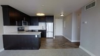 This single-floor space showcases a contemporary open-concept design that integrates the kitchen and living area seamlessly. The kitchen is appointed with dark espresso-toned cabinetry that extends to the ceiling, providing ample storage alongside a distinctive built-in wine rack above the refrigerator. A white subway tile backsplash creates a clean contrast against the dark cabinetry, while stainless steel appliances — including a French door refrigerator, electric range, and microwave — lend a polished, modern feel. A peninsula with a light-toned countertop extends into the space, offering additional prep and seating potential. Flush-mount ceiling lights provide consistent ambient illumination throughout. The kitchen and entry areas feature rich, dark hardwood-style flooring, which transitions to carpet in the adjacent living area, subtly defining each zone. The entryway is straightforward and functional, with neutral gray walls and clean white trim. Overall, the space presents a cohesive, modern aesthetic with a practical layout and a contrast-driven color palette of dark cabinetry, light walls, and mixed flooring materials.