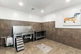 This purpose-built pet grooming wash room is located at 2370 Clark Fork Lane Rd in Missoula, Montana. The space is thoughtfully designed with both functionality and cleanliness in mind. Large-format, wood-toned ceramic tiles cover the floor and extend up the walls as full wainscoting, providing a durable, easy-to-clean surface throughout. The upper walls are finished in crisp white, creating a clean, bright contrast against the earthy tile tones.

The centerpiece of the room is a commercial-grade stainless steel dog wash tub, complete with an integrated handheld shower hose, plumbing connections, and a non-slip grated basin. A grooved step ramp alongside the tub allows pets of all sizes to enter and exit safely and comfortably. A stainless steel utility prep table with a lower shelf sits adjacent to the wash station, offering convenient storage and workspace for grooming supplies.

A central floor drain with a decorative mosaic stone surround ensures efficient water drainage, while recessed ceiling lighting and a ventilation grille contribute to a well-lit and properly ventilated environment. The entry features a sleek black-framed glass door, adding a modern architectural touch. Colorful, playful canvas prints of dogs adorn the walls, giving the space a welcoming and cheerful character suited to its purpose.