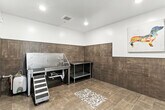 This purpose-built pet grooming wash room is located at 2370 Clark Fork Lane Rd in Missoula, Montana. The space is thoughtfully designed with both functionality and cleanliness in mind. Large-format, wood-toned ceramic tiles cover the floor and extend up the walls as full wainscoting, providing a durable, easy-to-clean surface throughout. The upper walls are finished in crisp white, creating a clean, bright contrast against the earthy tile tones.

The centerpiece of the room is a commercial-grade stainless steel dog wash tub, complete with an integrated handheld shower hose, plumbing connections, and a non-slip grated basin. A grooved step ramp alongside the tub allows pets of all sizes to enter and exit safely and comfortably. A stainless steel utility prep table with a lower shelf sits adjacent to the wash station, offering convenient storage and workspace for grooming supplies.

A central floor drain with a decorative mosaic stone surround ensures efficient water drainage, while recessed ceiling lighting and a ventilation grille contribute to a well-lit and properly ventilated environment. The entry features a sleek black-framed glass door, adding a modern architectural touch. Colorful, playful canvas prints of dogs adorn the walls, giving the space a welcoming and cheerful character suited to its purpose.