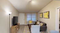 This single-floor space offers a comfortable and efficiently arranged living environment designed with both function and style in mind. The living area serves as the heart of the space, featuring warm beige walls, light wood-look flooring, and a textured ceiling that adds subtle character overhead. Track lighting provides adjustable illumination throughout the room, complementing the natural light that filters in through a sizable window dressed with horizontal blinds. The seating arrangement is welcoming and practical, centered around a gray sofa accented with decorative yellow and patterned throw pillows, a cushioned accent chair, and a cozy armchair, all anchored by a patterned area rug in gold and white tones. A rustic wooden media console hosts a large flat-screen television, adding a casual, lived-in warmth to the space. Bold abstract wall art in yellow and gray tones reinforces the cohesive color palette and adds visual interest to the walls. A white entry door with a sidelight window provides a clear focal point at the front of the room, making the layout feel open and easy to navigate. Overall, the space exudes a cheerful, inviting ambiance that balances comfort with contemporary design sensibilities.