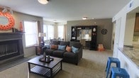 This single-floor space offers a thoughtfully designed open-concept layout that seamlessly integrates the living, dining, and work areas into one cohesive environment. The living area serves as the centerpiece, anchored by a plush dark charcoal sofa accented with vibrant teal and coral throw pillows, paired with a sturdy wooden coffee table that adds a grounded, earthy contrast. Soft carpeting throughout lends warmth and comfort underfoot. Natural light pours in generously through multiple windows dressed in neutral curtains, creating a bright and welcoming ambiance across the entire space. The walls are finished in a warm taupe tone that complements the furnishings and contributes to the overall cohesive aesthetic. A dark-finished shelving and entertainment unit along one wall provides ample storage and display space, functioning as a stylish focal point. Adjacent to the living area, a compact yet functional workspace is defined by a desk and task lighting, ideal for productivity. Further back, a dining area with a round table and chairs benefits from the same abundant natural light. The space strikes a balance between practical functionality and approachable, casual style, making it a comfortable and versatile environment suited for both relaxing and everyday living.