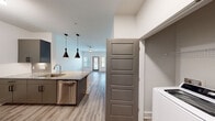 This well-appointed single-floor residence showcases a contemporary design with clean lines, neutral tones, and thoughtful finishes throughout. The open-concept layout connects the kitchen and living area seamlessly, creating a spacious and functional flow. The kitchen serves as the centerpiece, featuring granite countertops, a large island with an undermount sink, stainless steel appliances including a refrigerator, electric range, microwave, and dishwasher, as well as dark taupe cabinetry with modern hardware. A built-in wine rack and stylish pendant lighting with bold, sculptural fixtures add distinctive character to the space. Wood-look plank flooring runs throughout the entire residence, providing a warm yet easy-to-maintain surface. The living area is generously proportioned and bathed in natural light from multiple windows. A dedicated in-unit laundry area with a top-load washer is conveniently tucked off the kitchen corridor. Five-panel interior doors in a coordinating taupe finish lend a cohesive, polished aesthetic. The bedroom offers a bright, open feel with direct sightlines to the kitchen, while a hallway with additional storage connects the various zones of the home. The overall ambiance is modern and move-in ready, combining practical amenities with contemporary style.
