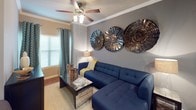 This thoughtfully designed single-floor residence offers a cohesive and stylish living experience throughout its open layout. Warm hardwood-style flooring flows through the main areas, complemented by neutral gray walls that create a versatile backdrop for the contemporary décor.

The living area is a welcoming focal point, featuring crown molding, a ceiling fan with light fixture, and a window dressed with teal patterned curtains that invite natural light. A bold navy sectional sofa anchors the space, while striking metallic spiral wall art adds a dramatic decorative element.

The galley-style kitchen is efficiently laid out with white cabinetry, dark granite countertops, stainless steel appliances including a full-size refrigerator and range, and a double basin sink. A dedicated laundry closet is conveniently accessible directly from the kitchen.

The bedroom is a serene retreat, finished with plush carpet and adorned with gold sunburst wall sculptures above the upholstered navy headboard. Twin nightstands flank the bed, and patterned drapes frame a bright window with shutters, enhancing the room's elegant character.

The bathroom offers a clean, well-lit vanity with white cabinetry, granite countertops, a large mirror, and hardwood-style flooring, maintaining the consistent design language found throughout the home.