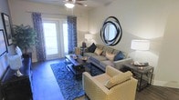 This single-floor space offers a thoughtfully designed living area that balances comfort and contemporary style. The living room serves as the centerpiece, featuring rich hardwood-style flooring that anchors the space with warmth and durability. A tufted gray sofa, patterned accent chair, and a plush navy area rug create a cohesive and inviting seating arrangement. A sleek coffee table with decorative accents adds functional elegance to the space.

A striking circular abstract wall mirror serves as a bold focal point above the sofa, complementing the neutral wall tones throughout. The room is illuminated by a combination of a ceiling fan with integrated lighting and stylish floor and table lamps, providing layered lighting options. Patterned drapes in deep blue and white frame the glass double doors, which invite natural light into the room while offering a connection to the outdoors. The overall ambiance is modern and well-curated, with a neutral color palette accented by deep blues and gold tones, resulting in a space that feels both polished and livable.