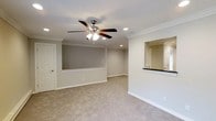 This single-floor space offers a clean, functional layout designed for comfortable everyday living. The floor plan is anchored by a spacious main living area finished with plush wall-to-wall carpeting and neutral gray walls that create a calm, welcoming atmosphere. Crown molding runs along the ceiling perimeter, lending a refined architectural touch, while recessed lighting and a ceiling fan with a light fixture ensure the room is well-lit and climate-friendly year-round. A pass-through window connects the living area to the kitchen, enhancing openness and communication between spaces. The kitchen features dark countertops and is positioned centrally within the layout, offering convenient access to both sides of the floor plan. Natural light filters in through windows on opposing ends of the space, contributing to a bright and airy feel throughout. The second main room mirrors the overall design language, maintaining consistent carpeting and neutral tones that unify the space. White interior doors and crisp white trim throughout further reinforce the clean, cohesive aesthetic. Overall, the space presents a straightforward yet polished interior ideal for those seeking a comfortable and move-in-ready environment.