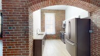 This single-floor unit showcases a compelling combination of historic industrial character and contemporary updates. The space is defined by prominent exposed red brick walls throughout, providing a warm, textured backdrop that contrasts beautifully with clean white walls and light-toned hardwood-style flooring. Structural steel columns and exposed ductwork run overhead, reinforcing the loft's industrial aesthetic.

The open living area benefits from generous natural light streaming through large multi-pane windows dressed with sheer curtains. A striking barrel-vaulted brick archway serves as a dramatic architectural transition into the kitchen, which features two-tone cabinetry pairing dark wood lower cabinets with white uppers, quartz countertops, a white subway tile backsplash, and a full suite of stainless steel appliances including a refrigerator, range, and over-the-range microwave.

The bedroom offers wood-look flooring, exposed ductwork, and a partial brick accent wall with a window. The bathroom is cleanly finished with large-format tile flooring, a wall-mounted floating vanity with an illuminated mirror, and a glass-enclosed walk-in shower. An in-unit stacked washer and dryer is also present, adding everyday convenience to this character-rich space.