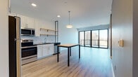 Located at 237 S Second St in Milwaukee's vibrant Walker's Point neighborhood, this single-floor unit offers a thoughtfully designed open-concept layout with contemporary finishes throughout. The kitchen is a standout feature, equipped with stainless steel appliances including a range, over-the-range microwave, refrigerator, and dishwasher. White shaker-style cabinetry pairs cleanly with dark countertops and a classic white subway tile backsplash, while open wood shelving adds a modern industrial touch. Light wood-style flooring extends seamlessly from the kitchen into the expansive living and dining area, where a sleek pendant light fixture anchors the space. Floor-to-ceiling windows with black metal frames flood the living area with natural light and open to a private balcony with elevated views of the Milwaukee cityscape. The bedroom features plush carpeting and a generous built-in closet with wire shelving for ample storage. The bathroom is finished with a white vanity, dark countertop, and a large mirror with overhead lighting. A dedicated laundry area accommodates a stacked full-size washer and dryer, adding everyday convenience. The overall design balances clean, modern aesthetics with functional, well-appointed living spaces.