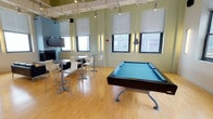 This single-floor amenity space is designed as a multifunctional lounge and recreation area, combining social, leisure, and casual gathering zones within an open, airy layout. The interior is characterized by high ceilings with exposed industrial ductwork and track lighting, light hardwood flooring throughout, and a vibrant color palette of sage green, sky blue, and crisp white that gives the space a modern, energetic feel.

The main lounge area features black leather seating arranged around a minimalist white coffee table, complemented by a striking blue accent column housing a fireplace with a wall-mounted flat-screen TV above it. Vertical LED wall sconces add warm ambient lighting alongside the large roller-shaded windows, which frame views of the surrounding urban cityscape and fill the space with natural light.

Adjacent to the lounge, a recreation zone includes a full-size pool table with a teal felt surface, while high-top tables and bar-height chairs provide casual seating for socializing or informal gatherings. The open corridor area, defined by bold blue structural columns, connects the zones seamlessly.

A dedicated kitchen area features blue cabinetry, white countertops, a sink, stainless steel appliances including a beverage cooler, and a wall-mounted TV, making it well-equipped for entertaining and events.