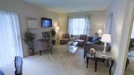 This single-floor living space offers a thoughtfully designed interior centered around a spacious, open living area. The room features soft, neutral wall tones complemented by plush wall-to-wall carpeting, creating a warm and inviting atmosphere. A wall-mounted flat-screen television serves as a focal point, flanked by decorative artwork and a sleek console. Natural light flows in through a large central window dressed with elegant patterned curtains and blinds, brightening the space throughout the day. The seating arrangement includes a mix of patterned accent chairs and a bold zebra-print sofa, adding visual interest while maintaining a cohesive design aesthetic. A mirrored glass coffee table anchors the conversation area, reflecting light and contributing to the room's airy feel. Decorative potted plants are strategically placed throughout, introducing organic texture and color. Additional floor lamps provide warm ambient lighting for evening use. The overall ambiance is polished yet comfortable, blending contemporary style with practical living elements in a well-proportioned, single-level layout.