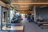 Located at 2333 Calle Del Mundo in Santa Clara, CA, this professional-grade fitness center offers a generously sized, single-floor exercise facility designed to accommodate a wide range of workout needs. The space features an industrial-modern aesthetic, with exposed ductwork, structural columns, and warm linear LED lighting suspended from an open ceiling — creating a clean, energetic atmosphere throughout.

The facility is divided into distinct training zones. A dedicated cardio area lines the perimeter with large floor-to-ceiling windows, flooding the space with natural light and providing open outdoor views. Multiple treadmills, ellipticals, stair climbers, and stationary bikes — primarily Matrix-branded — are thoughtfully arranged for comfortable use. A strength training zone features cable machines, adjustable benches, a power rack with Olympic plates, and a comprehensive dumbbell rack.

A separate functional training area includes open floor space ideal for dynamic movement, complemented by a cable functional trainer, plyo boxes, and additional free weights. Durable rubber flooring throughout provides traction and impact absorption suited for high-intensity use. The entrance area features patterned accent walls and glass-panel doors, adding a polished, welcoming touch. A water station is also present for user convenience. Overall, the space is well-equipped, well-lit, and thoughtfully laid out for diverse fitness goals.
